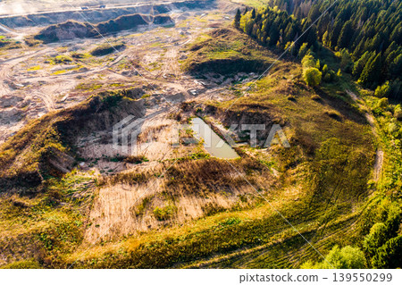 Aerial view of an open-pit sand quarry featuring excavated terrain, dirt tracks, and a small pool. Lush green forest edges the active mining site, showcasing industrial impact on nature Aerial view of an open-pit sand quarry featuring excavated terrain, dirt tracks, and a small pool. Lush green forest edges the active mining site, showcasing industrial impact on nature 139550299