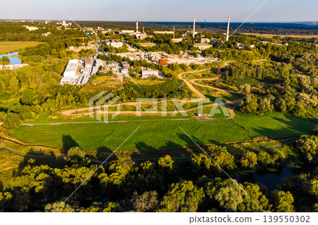Drone shot of a bustling industrial complex with towering stacks, surrounded by verdant fields and a meandering river, bathed in warm golden light Drone shot of a bustling industrial complex with towering stacks, surrounded by verdant fields and a meandering river, bathed in warm golden light 139550302