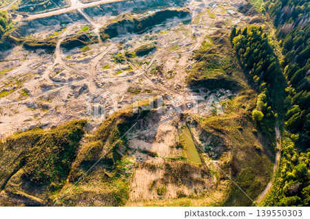 Dynamic aerial view of an active open-pit quarry, showcasing vast sandy excavations with numerous vehicle tracks. A small pond contrasts with the golden earth, bordering a dense pine forest 139550303