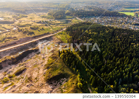 Aerial perspective of a new active sand quarry alongside a verdant forest and a distant village, starkly highlighting human land alteration Aerial perspective of a new active sand quarry alongside a verdant forest and a distant village, starkly highlighting human land alteration 139550304