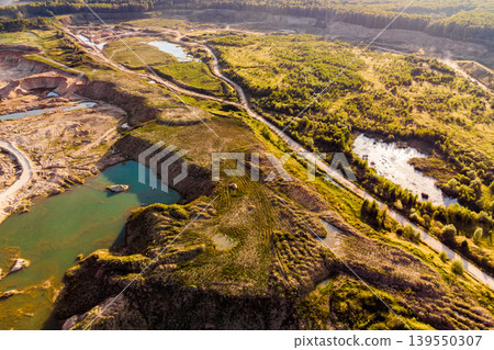 Aerial view of a vast open-pit quarry with turquoise water bodies and rugged terrain. Dirt roads snake through the landscape, bordered by lush green vegetation. Industrial extraction meets natural Aerial view of a vast open-pit quarry with turquoise water bodies and rugged terrain. Dirt roads snake through the landscape, bordered by lush green vegetation. Industrial extraction meets natural 139550307