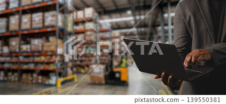 Worker using laptop in modern warehouse with storage shelves and cargo boxes, suitable for smart warehouse management system, logistics monitoring, inventory tracking, and industrial supply 139550381