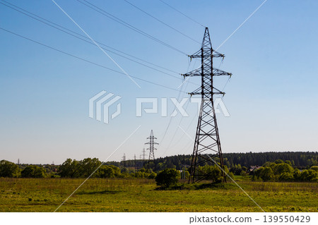 Imposing metal power line tower stands tall in a lush green field. Its robust structure supports numerous high-voltage lines against a clear sky 139550429