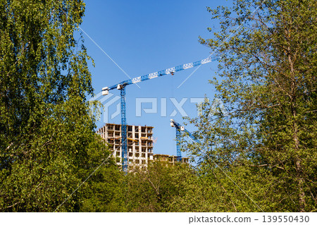 Tower cranes busy on a high-rise construction site, structure rising amidst green trees under a clear blue sky Tower cranes busy on a high-rise construction site, structure rising amidst green trees under a clear blue sky 139550430