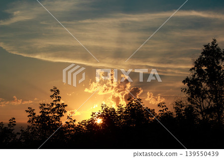 Fiery sunset peeking through silhouetted trees, painting the sky with warm hues. Dramatic clouds catch golden sunbeams at dusk, a tranquil nature scene Fiery sunset peeking through silhouetted trees, painting the sky with warm hues. Dramatic clouds catch golden sunbeams at dusk, a tranquil nature scene 139550439