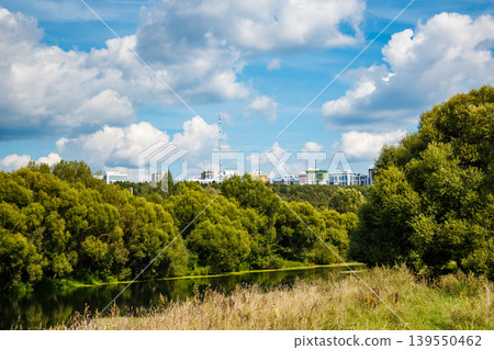 Green river flanked by lush trees, tranquil waters reflect the clear sky. Distant city buildings and a tower peek over the dense foliage under cumulus clouds Green river flanked by lush trees, tranquil waters reflect the clear sky. Distant city buildings and a tower peek over the dense foliage under cumulus clouds 139550462