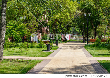 Inviting paved park alley on a bright sunny day, flanked by vibrant green trees, lush grass, benches, and streetlights. A serene natural escape Inviting paved park alley on a bright sunny day, flanked by vibrant green trees, lush grass, benches, and streetlights. A serene natural escape 139550475