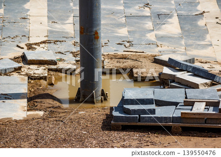 Rusty metal pillar stands tall in a grimy puddle, amidst scattered, broken paving stones. Construction site mess with dislodged pavers 139550476