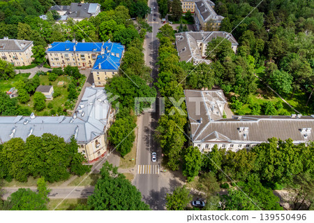 Aerial shot reveals a tree-lined city street, a white car on a crosswalk, bordered by old-school residential buildings. Green foliage pops Aerial shot reveals a tree-lined city street, a white car on a crosswalk, bordered by old-school residential buildings. Green foliage pops 139550496