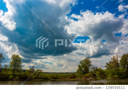 Majestic stormy clouds dominate, some fluffy white, others brooding grey, with vivid blue sky peeking through. Lush riverbanks and trees below Majestic stormy clouds dominate, some fluffy white, others brooding grey, with vivid blue sky peeking through. Lush riverbanks and trees below 139550511
