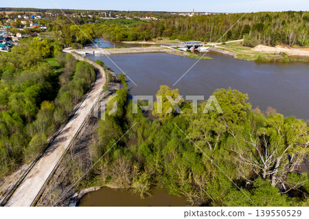 Aerial view of a scenic river dam and sluice gates, flanked by vibrant spring foliage. A rugged path hugs the calm, brownish waters, with distant village dwellings peeking through Aerial view of a scenic river dam and sluice gates, flanked by vibrant spring foliage. A rugged path hugs the calm, brownish waters, with distant village dwellings peeking through 139550529