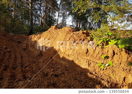Earthen trench with a freshly dug mound of soil, illuminated by sunlight. Trees in the background offer a natural backdrop, highlighting construction or landscaping 139550834