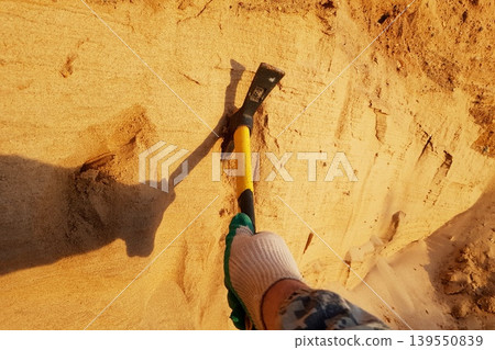 A gloved hand expertly wields a bright pickaxe, carving into sunlit sandy earth. The tool's shadow stretches, revealing diligent excavation efforts A gloved hand expertly wields a bright pickaxe, carving into sunlit sandy earth. The tool's shadow stretches, revealing diligent excavation efforts 139550839