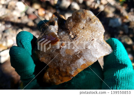 Gloved hand grips a stunning geode specimen, featuring shimmering drusy quartz crystals. Its rugged, earthy surface catches the sun, blurring the rocky background Gloved hand grips a stunning geode specimen, featuring shimmering drusy quartz crystals. Its rugged, earthy surface catches the sun, blurring the rocky background 139550853