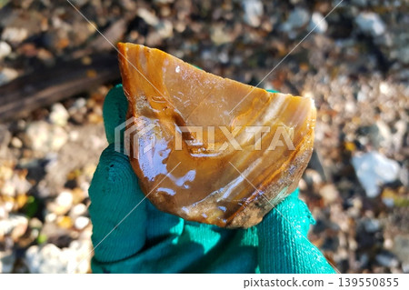A gloved hand holds a slick, banded rock piece. Its rich amber and sandy tones shimmer, revealing cool natural striations under bright light 139550855