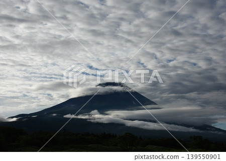 Lenticular clouds cut in front of the dark Mount Fuji, and large, cirrocumulus clouds seem to rise from behind. 139550901
