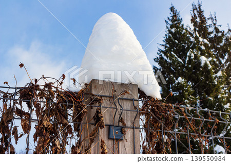 Fluffy snow crowns a rustic wooden fence post, intricately entwined with dry winter vines under a crisp blue sky, a stark natural contrast Fluffy snow crowns a rustic wooden fence post, intricately entwined with dry winter vines under a crisp blue sky, a stark natural contrast 139550984