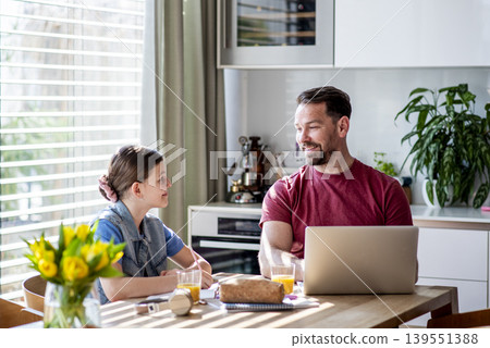 Daughter studying at table as dad works remotely on laptop nearby. 139551388