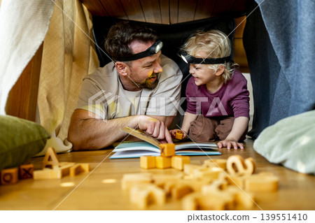 Dad and child building and reading book in cozy blanket fort. Dad and child building and reading book in cozy blanket fort. 139551410