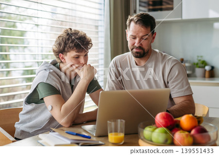 Father helping teenage son with diabetes with homework on laptop in kitchen. 139551433