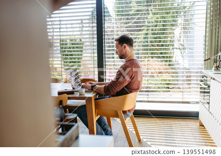 Man sitting in kitchen working remotely on laptop. Man sitting in kitchen working remotely on laptop. 139551498