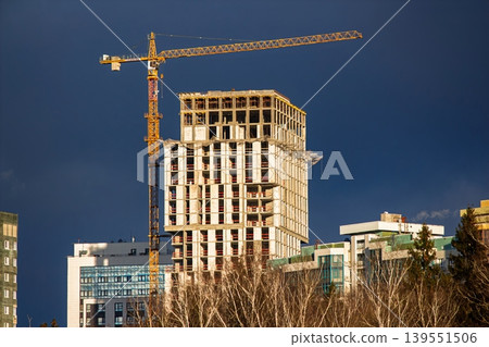 Tower crane and contemporary high-rise building under construction, reflecting dynamic urban development. Dramatic dark sky backdrop emphasizes scale and ambition of city growth Tower crane and contemporary high-rise building under construction, reflecting dynamic urban development. Dramatic dark sky backdrop emphasizes scale and ambition of city growth 139551506