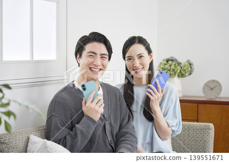 A portrait of a couple in their living room, holding a smartphone and looking directly at the camera. 139551671