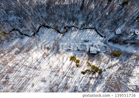 Aerial view of a winding creek cutting through snow-covered winter forest with isolated cabins and long shadows Aerial view of a winding creek cutting through snow-covered winter forest with isolated cabins and long shadows 139551716