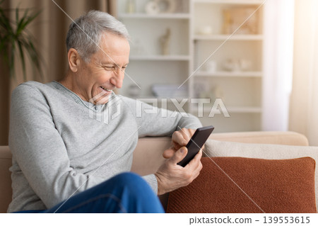Cheerful mature man using smartphone while sitting on sofa at home Cheerful mature man using smartphone while sitting on sofa at home 139553615