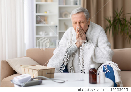 Sick senior man sneezing into tissue while wrapped in blanket at home 139553655