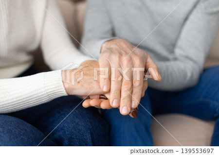 Closeup of two mature people holding hands while sitting together at home Closeup of two mature people holding hands while sitting together at home 139553697