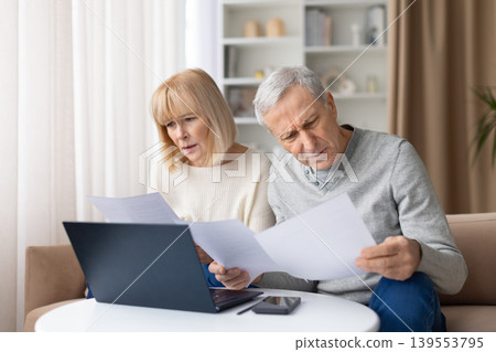 Concerned senior couple reading financial papers at home, looking worried Concerned senior couple reading financial papers at home, looking worried 139553795