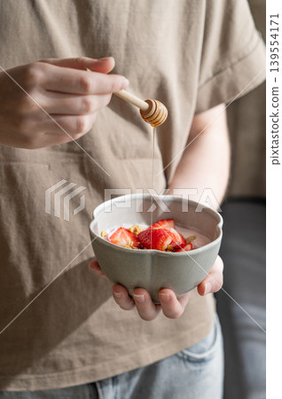 A person holding a ceramic bowl of yogurt, fresh strawberries, and granola, drizzling with honey. A person holding a ceramic bowl of yogurt, fresh strawberries, and granola, drizzling with honey. 139554171