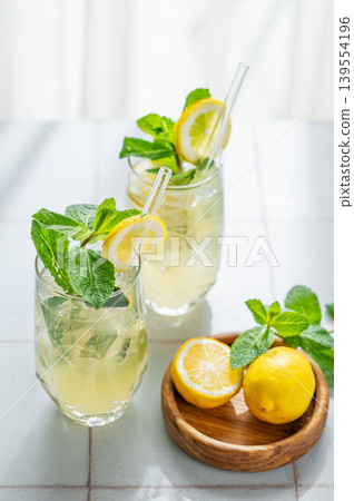 A summer drink with lemon, mint, and ice in a two glasses against a sunny window background. A summer drink with lemon, mint, and ice in a two glasses against a sunny window background. 139554196