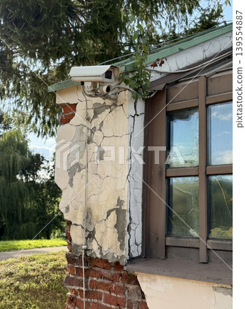 Security camera mounted on weathered building corner amidst greenery and cracked walls Security camera mounted on weathered building corner amidst greenery and cracked walls 139554887