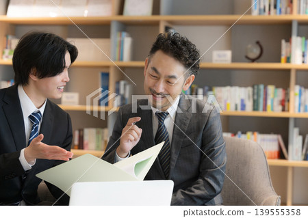 A client's CEO and two young businessmen are having a meeting on laptops with a bookshelf in the background, discussing proposals and explanations for corporate sales. A client's CEO and two young businessmen are having a meeting on laptops with a bookshelf in the background, discussing proposals and explanations for corporate sales. 139555358