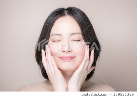 A close-up image of a smiling woman with her eyes closed, touching her translucent, beautiful skin against a beige background, showcasing skincare and beauty. A close-up image of a smiling woman with her eyes closed, touching her translucent, beautiful skin against a beige background, showcasing skincare and beauty. 139555359