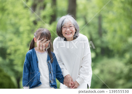 A grandmother holding hands with her granddaughter, who is smiling and looking at the camera, as they play in a park surrounded by the fresh greenery of spring. A grandmother holding hands with her granddaughter, who is smiling and looking at the camera, as they play in a park surrounded by the fresh greenery of spring. 139555530