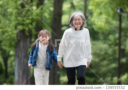 A grandmother holding hands with her granddaughter, who is smiling and looking at the camera, as they play in a park surrounded by the fresh greenery of spring. A grandmother holding hands with her granddaughter, who is smiling and looking at the camera, as they play in a park surrounded by the fresh greenery of spring. 139555532