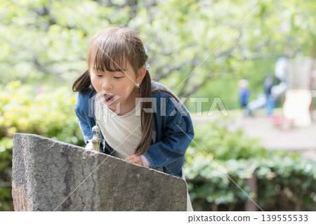 A portrait of a girl rehydrating at a water fountain in a park; an image of a hot day, with fresh greenery. A portrait of a girl rehydrating at a water fountain in a park; an image of a hot day, with fresh greenery. 139555533