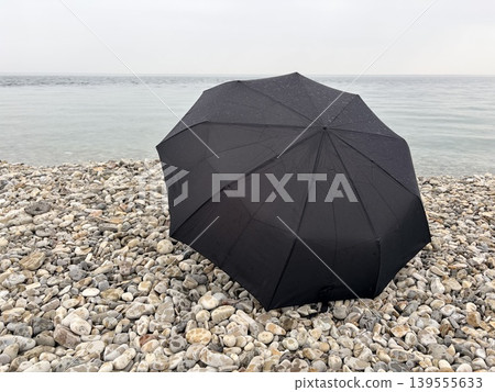 a minimalist, serene seascape featuring a large black umbrella resting open on a pebble beach. a minimalist, serene seascape featuring a large black umbrella resting open on a pebble beach. 139555633