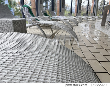 a low-angle, close-up perspective of a row of white wicker sun loungers arranged along the curved edge of an indoor pool or spa area. a low-angle, close-up perspective of a row of white wicker sun loungers arranged along the curved edge of an indoor pool or spa area. 139555655
