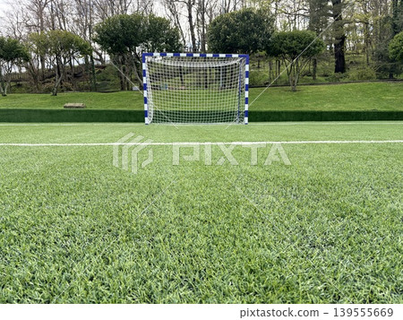 a bright, well-maintained soccer field covered in vibrant green artificial turf. Soccer goal featuring a white net and posts painted with blue and white vertical stripes. a bright, well-maintained soccer field covered in vibrant green artificial turf. Soccer goal featuring a white net and posts painted with blue and white vertical stripes. 139555669