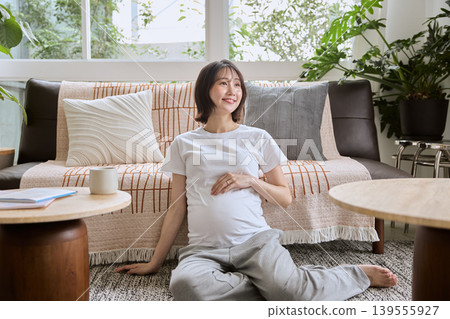 A pregnant woman sits on the living room floor with her hand on her stomach, smiling. A pregnant woman sits on the living room floor with her hand on her stomach, smiling. 139555927