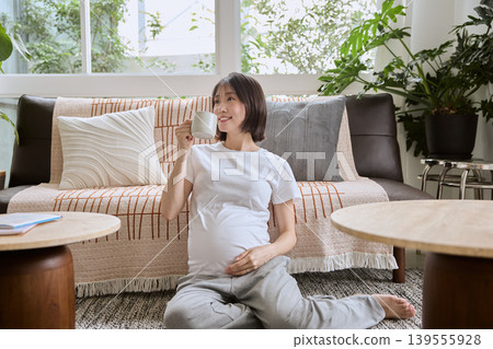 一位孕婦坐在客廳地板上,手裡拿著杯子,面帶微笑。 一位孕婦坐在客廳地板上,手裡拿著杯子,面帶微笑。 139555928