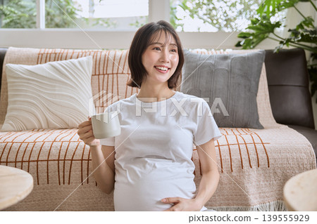 A smiling pregnant woman relaxing on the living room sofa, holding a mug. A smiling pregnant woman relaxing on the living room sofa, holding a mug. 139555929