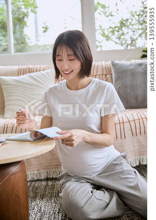 A pregnant woman smiles as she writes in her maternal and child health handbook in the living room. 139555935