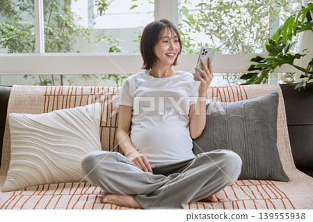 A pregnant woman smiles while looking at her smartphone on the sofa. 139555938