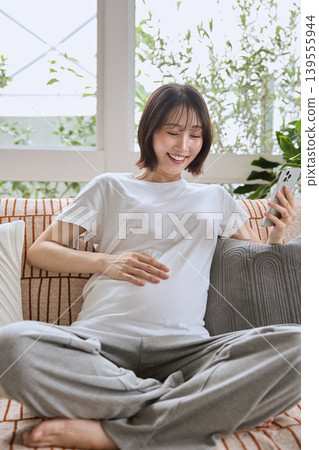 A pregnant woman smiles and touches her belly while looking at her smartphone on the sofa. A pregnant woman smiles and touches her belly while looking at her smartphone on the sofa. 139555944