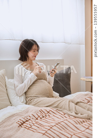 A young woman with a smile sits on a bed and operates her smartphone. A young woman with a smile sits on a bed and operates her smartphone. 139555957